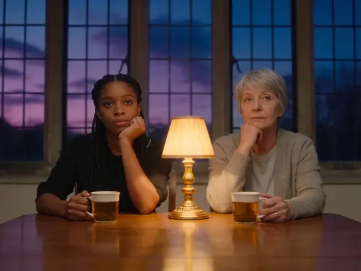 A young Black woman and an older white woman sit at a long table by tall windows at dusk, each with a mug, thinking.