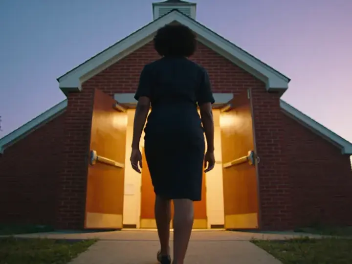 A Black woman walks up the path towards the open, lit doors of a red-brick hall at dusk.