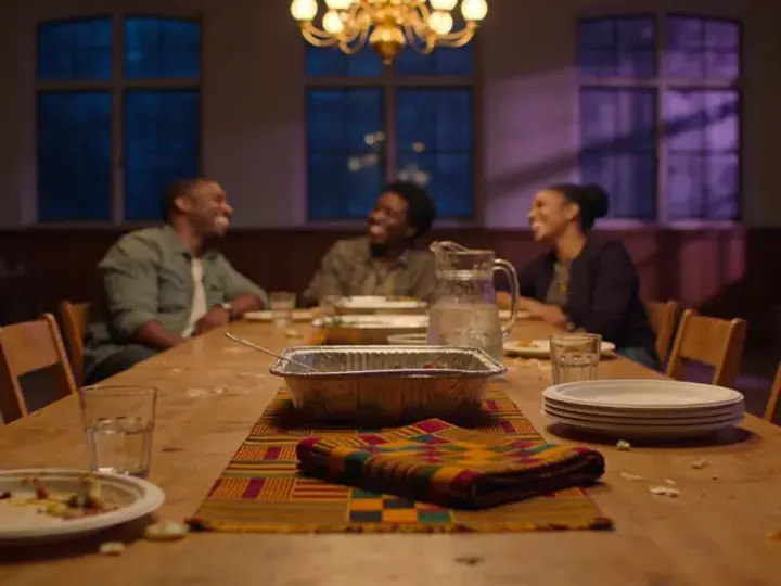 Three Black people laugh together around a long table after a shared meal, a kente cloth across the middle.